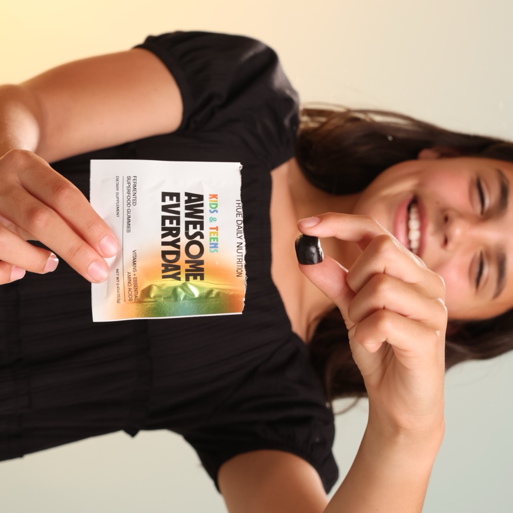 A smiling kid holding an Awesome Everyday packet and a gummy