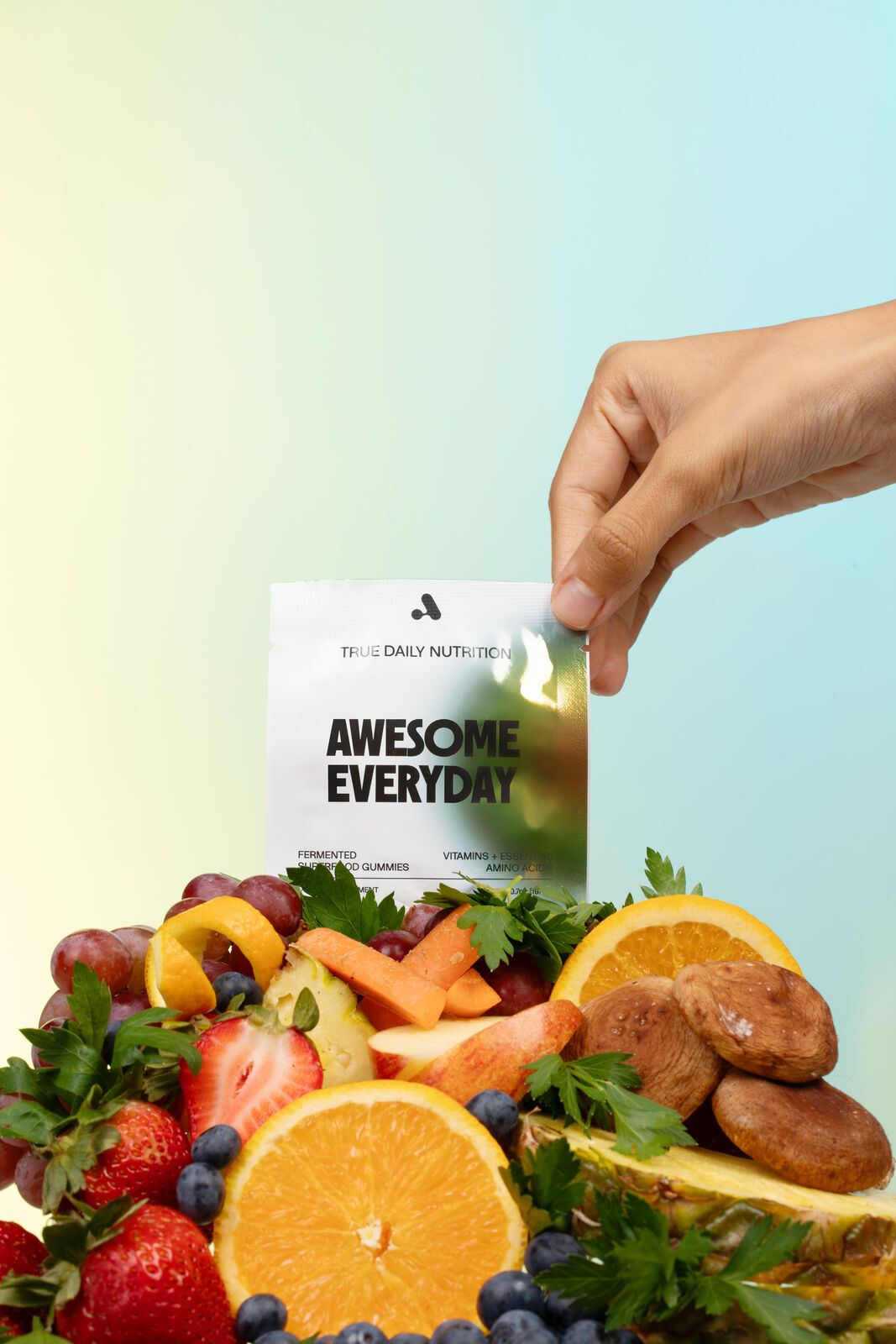 A hand holding the Awesome Everyday pack above fresh fruits and superfoods